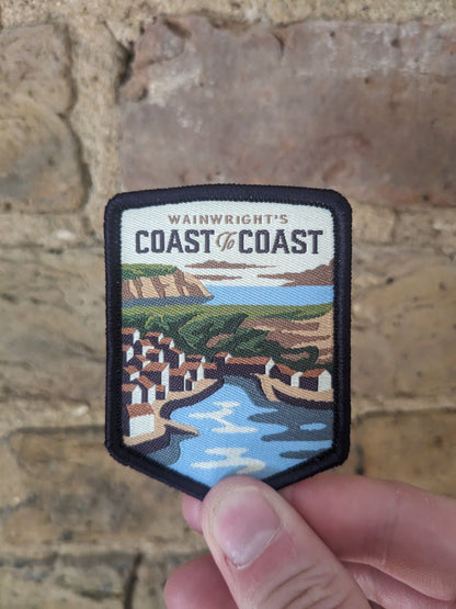 Image of the coast to coast patch being held against the backdrop of a brick wall. The patch depicts the Robin Hood's Bay, classic ending point of the Coast to Coast walk if walked west to east.