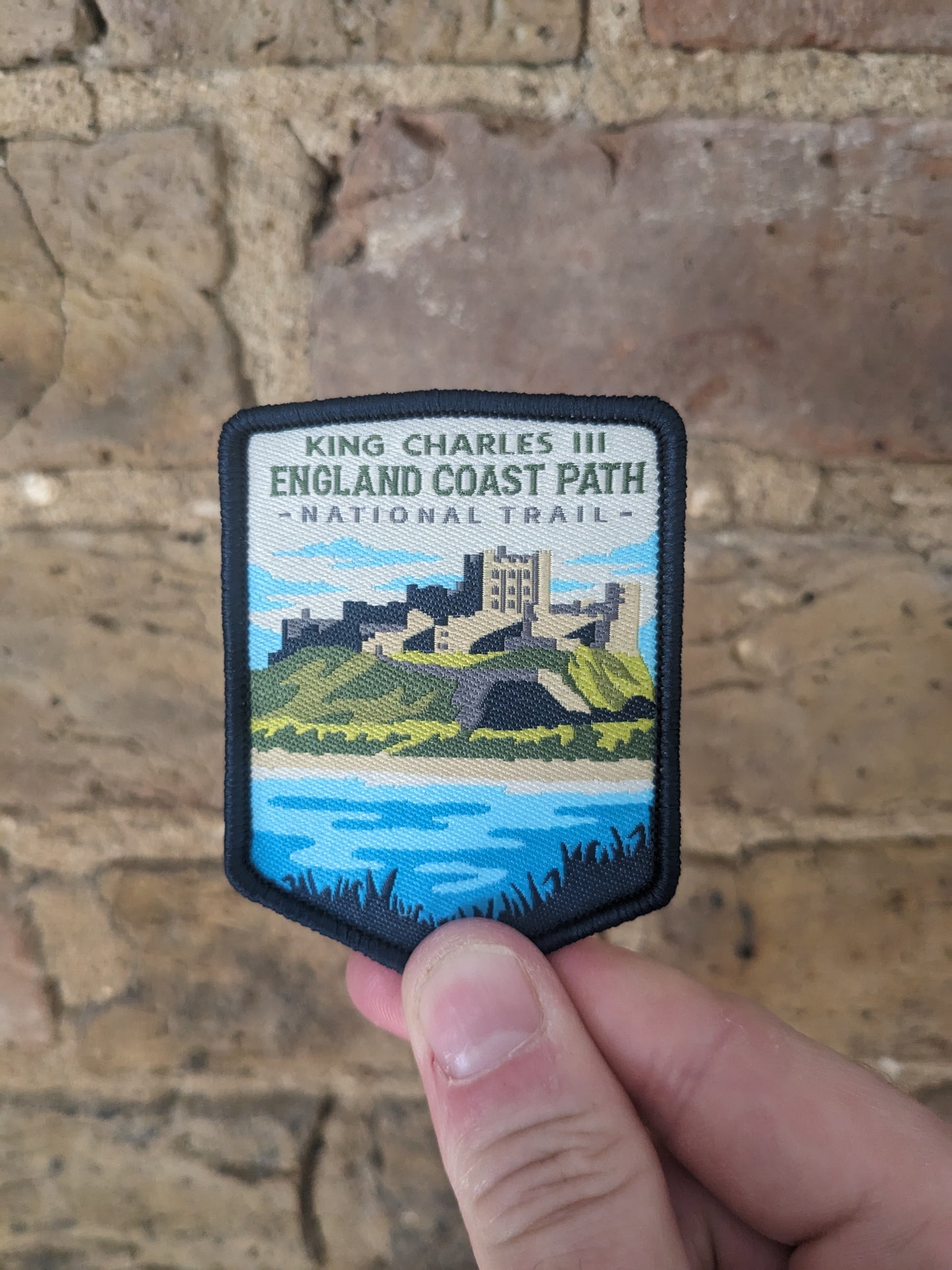 A badge/patch of the England Coast Path being held up against a brick wall backdrop. The patch itself features a view of Bamburgh Castle.