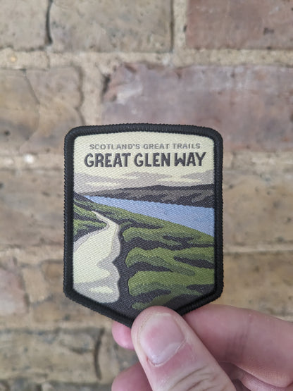 Image holding the Great Glen Way badge / patch against a backdrop of a brick wall. The patch itself features a view overlooking Loch Ness from the trail.