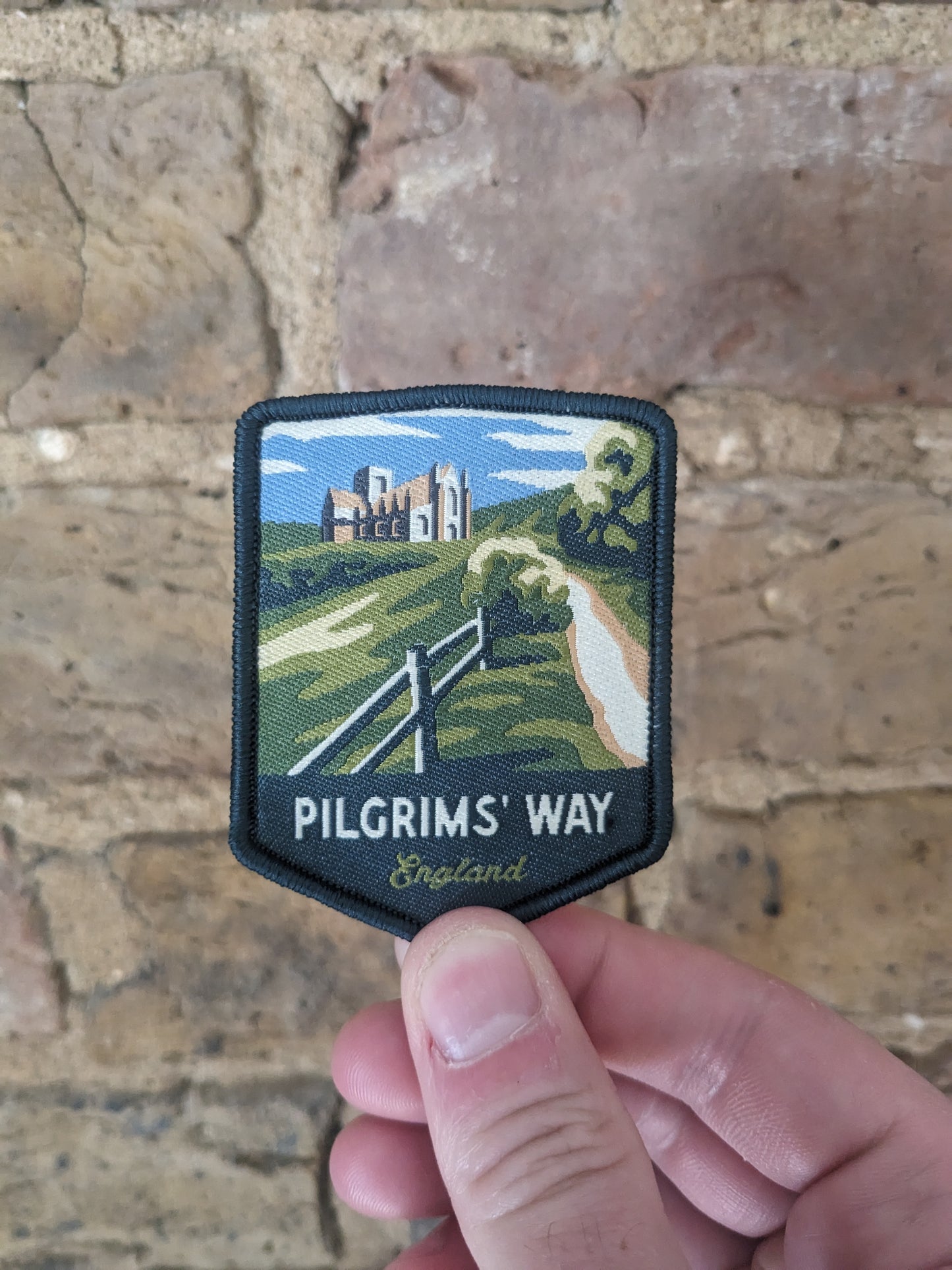 Image showing the Pilgrims' Way patch being held against a brick wall. The patch depicts Winchester Cathedral surrounded by bucolic countryside.