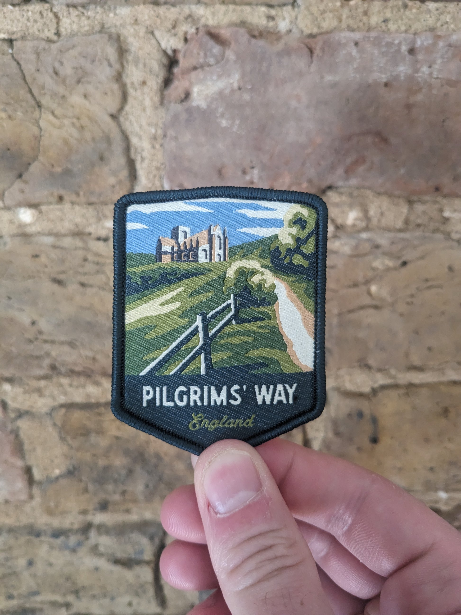 Image showing the Pilgrims' Way patch being held against a brick wall. The patch depicts Winchester Cathedral surrounded by bucolic countryside.