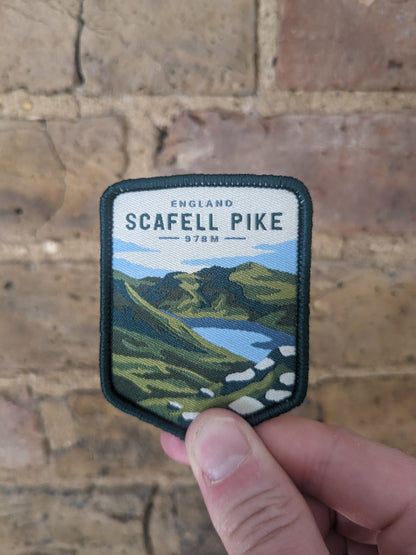 Image holding the Scafell Pike patch against a brick wall backdrop. The badge features the iconic mountain in the distance.