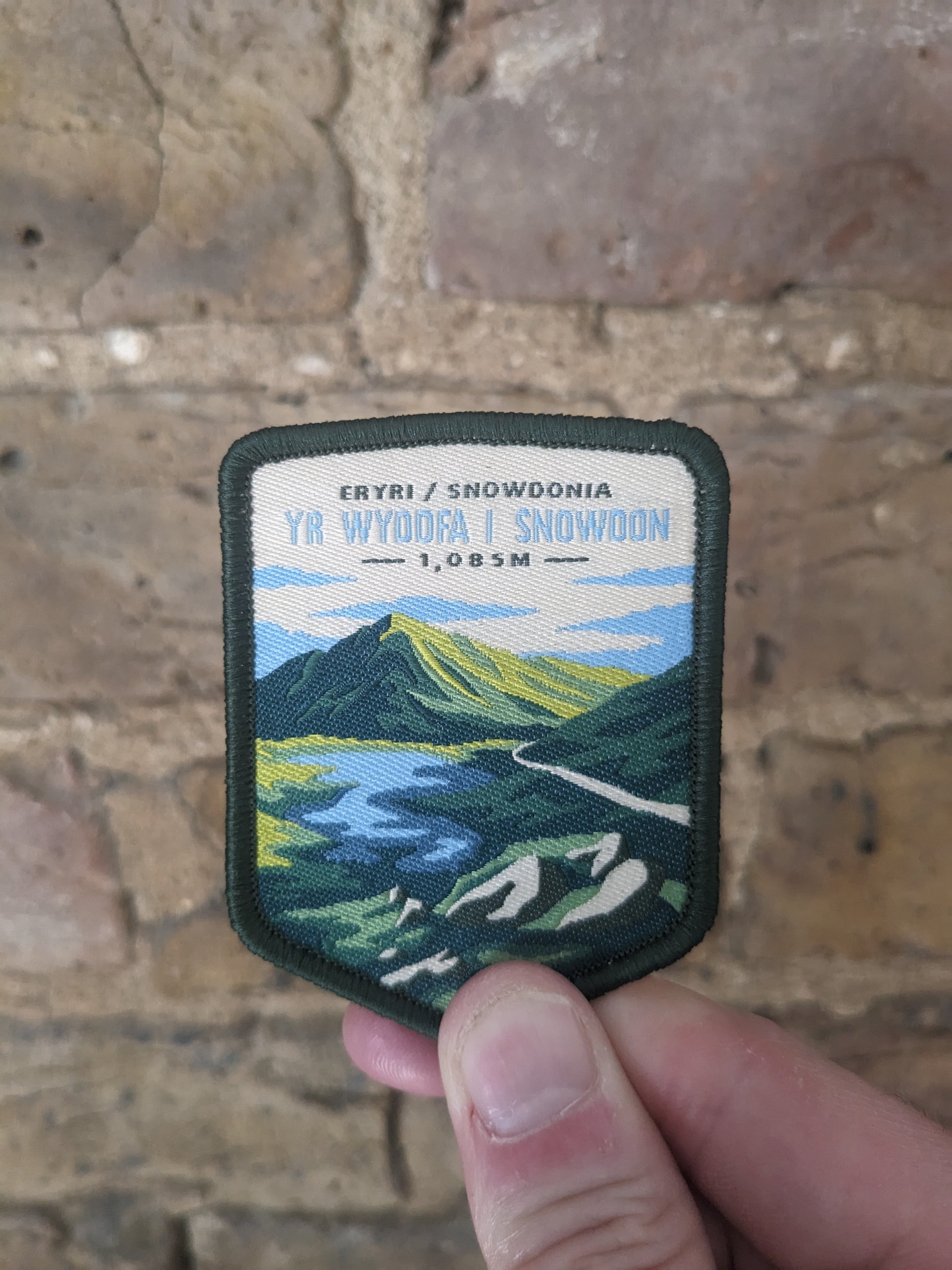 Image of the Yr Wyddfa / Snowdon patch being held against a backdrop of a brick wall. The badge itself features the iconic mountain from a distance.