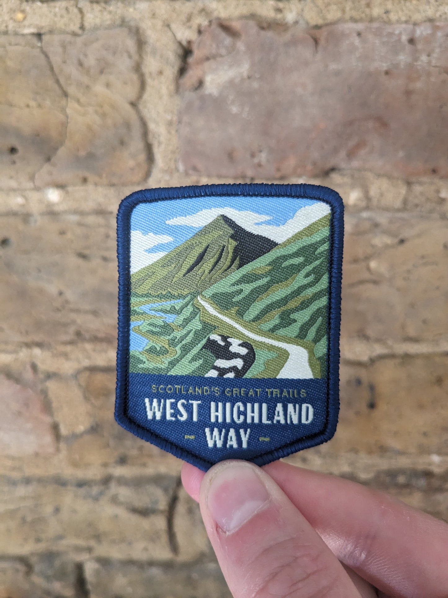 Picture of the West Highland Way badge being held up against a brick wall backdrop